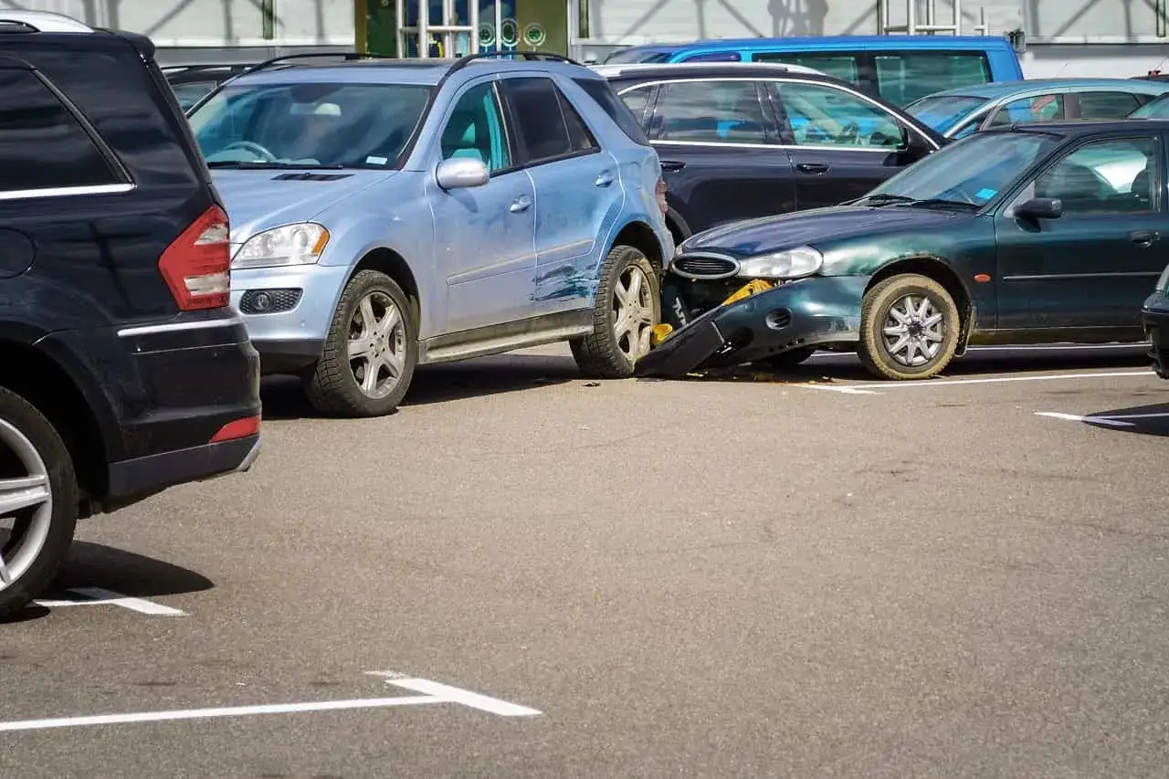 Na parkingu stoi kilka samochodów, jeden z nich ma uszkodzony zderzak. To ewidentna szkoda parkingowa.
