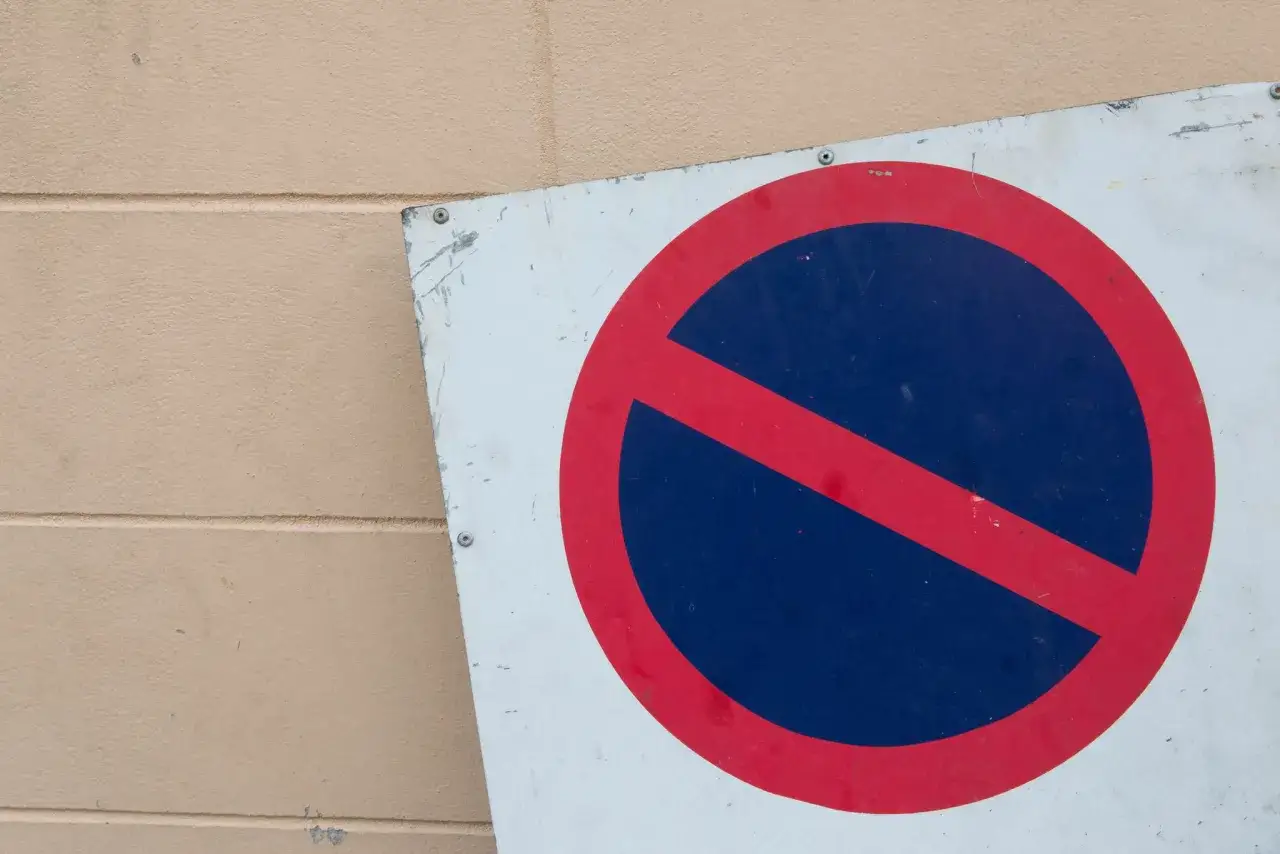 Znak zakaz postoju na białej tablicy, przytwierdzonej do jasnobrązowej ściany.
