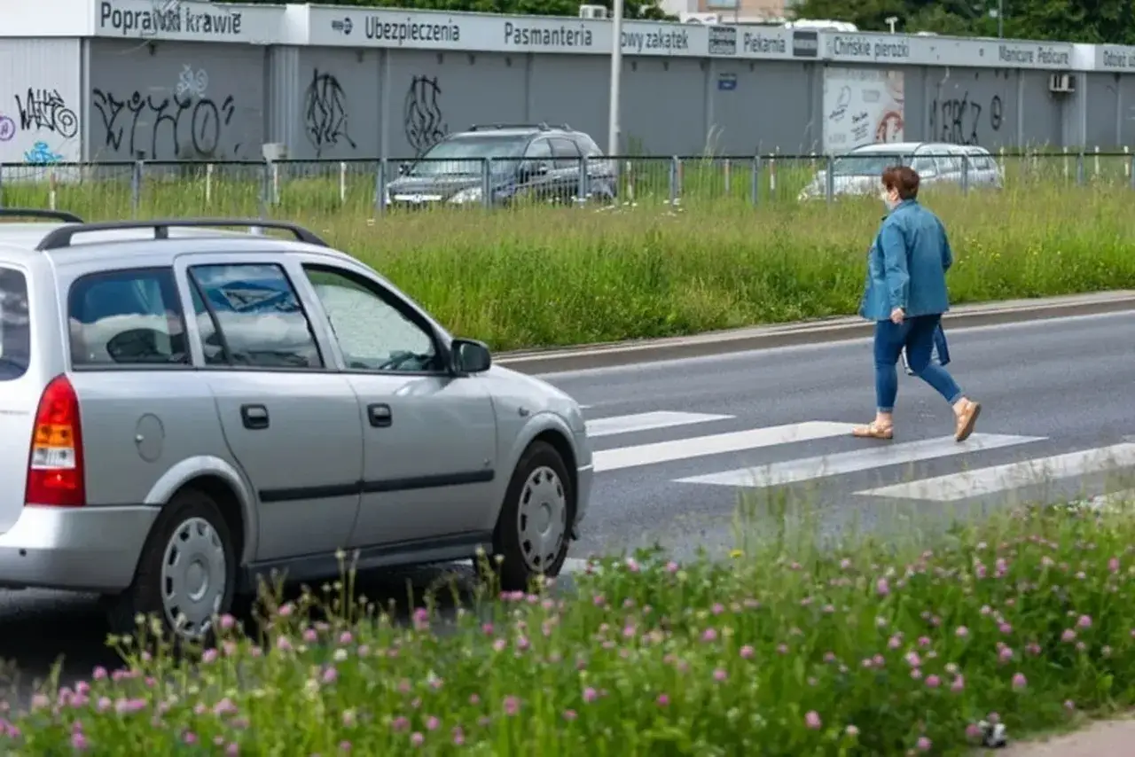 Kobieta w dżinsowej kurtce przechodzi przez pasy, kiedy pieszy ma pierwszeństwo. Srebrny samochód czeka.
