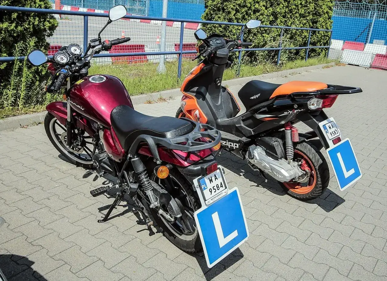 Dwa pojazdy, motorower i skuter, gotowe do nauki jazdy. Na każdym z nich widnieje tablica z literą