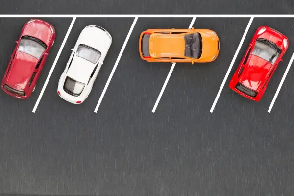 Czerwony, biały i pomarańczowy samochodzik na parkingu. Wykonują parkowanie równoległe przodem, zajmując miejsca wyznaczone białymi liniami.